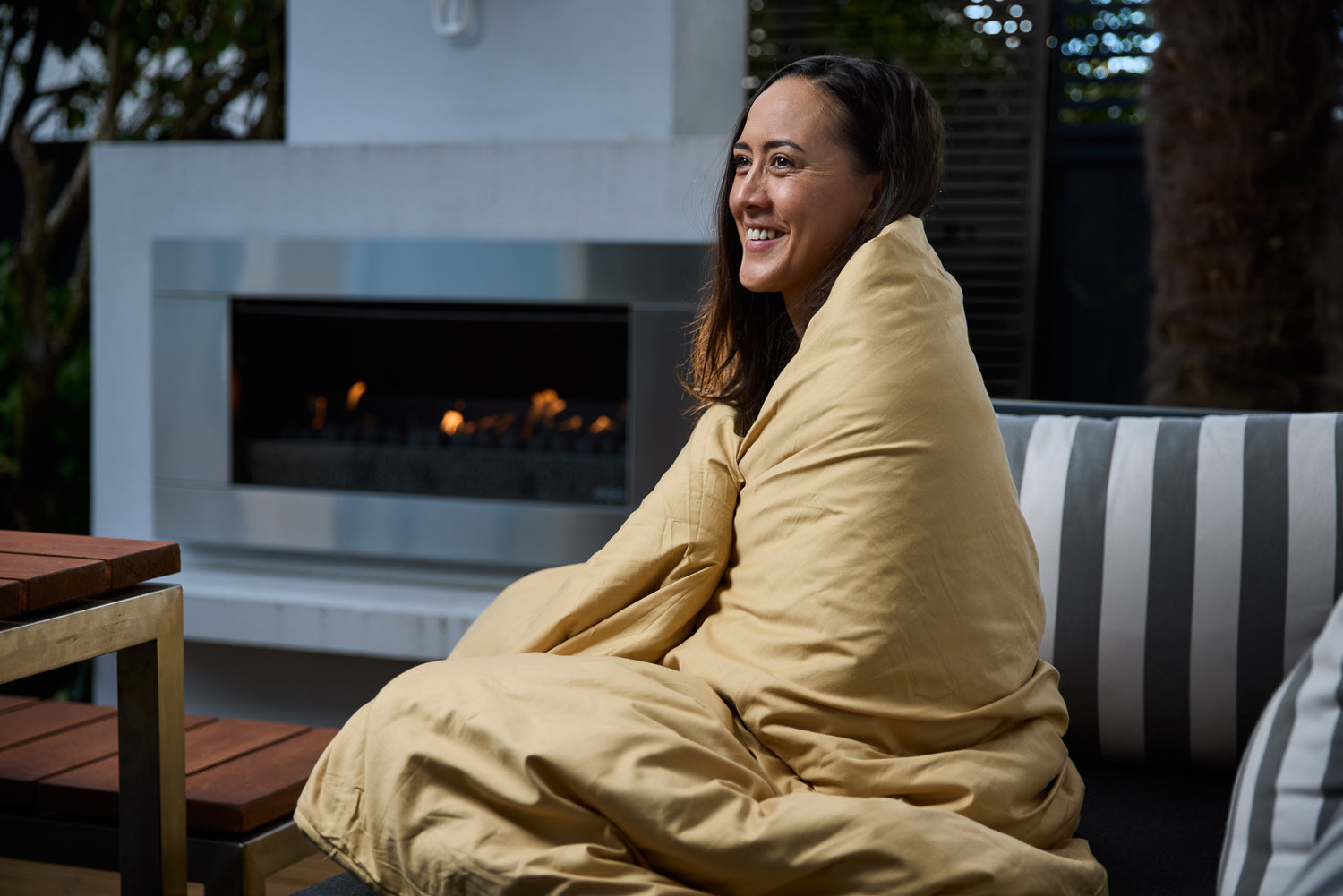 Woman outside wrapped in the Somni premium weighted blanket with an open fire behind her